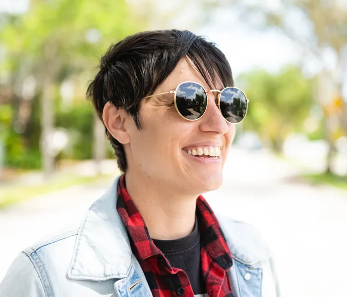 gender neutral person smiling and wearing a denim jacket and sunglasses on a sunny day